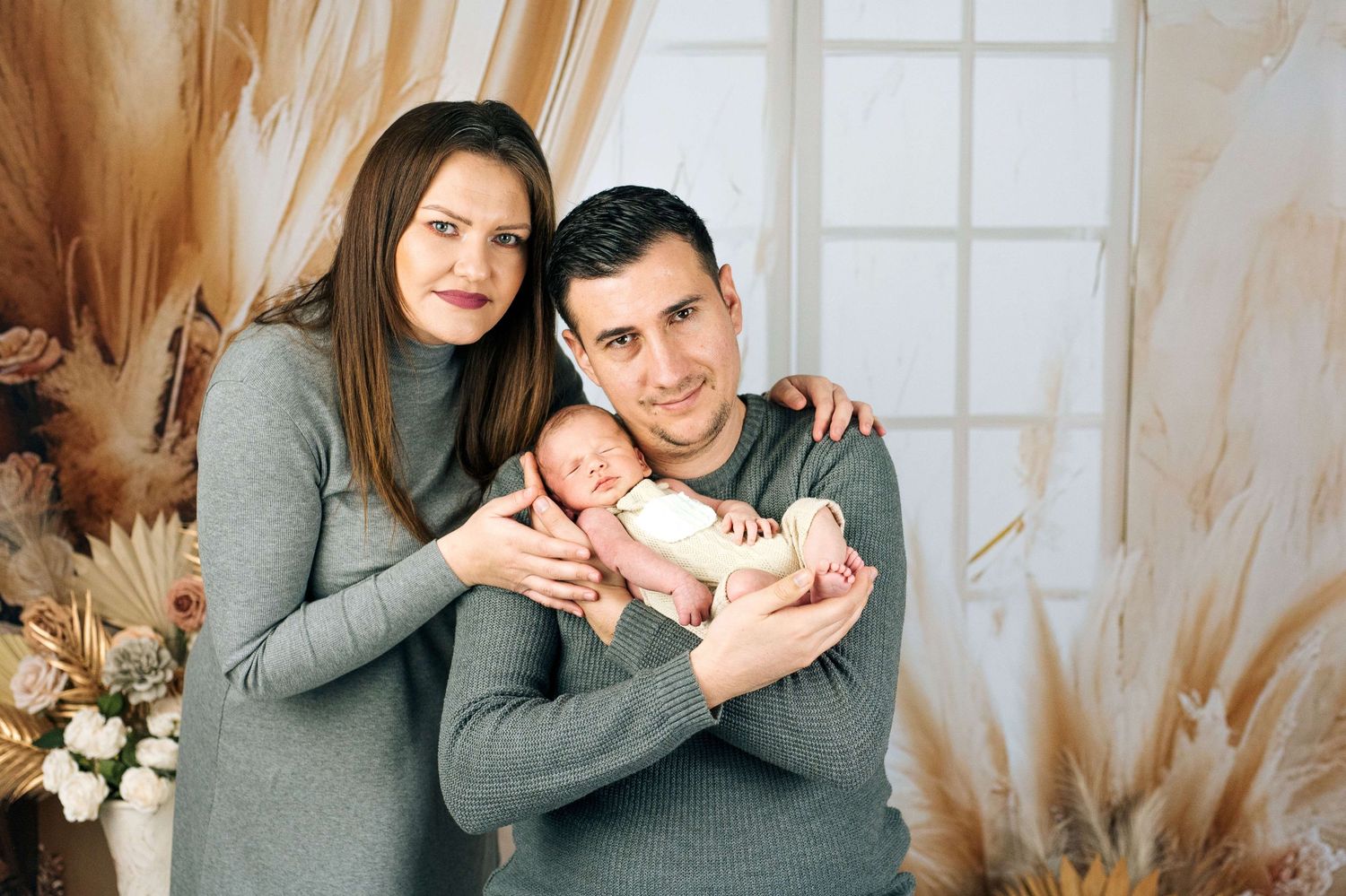 A happy couple in matching gray outfits holding their newborn baby in a cozy decorated studio setting.