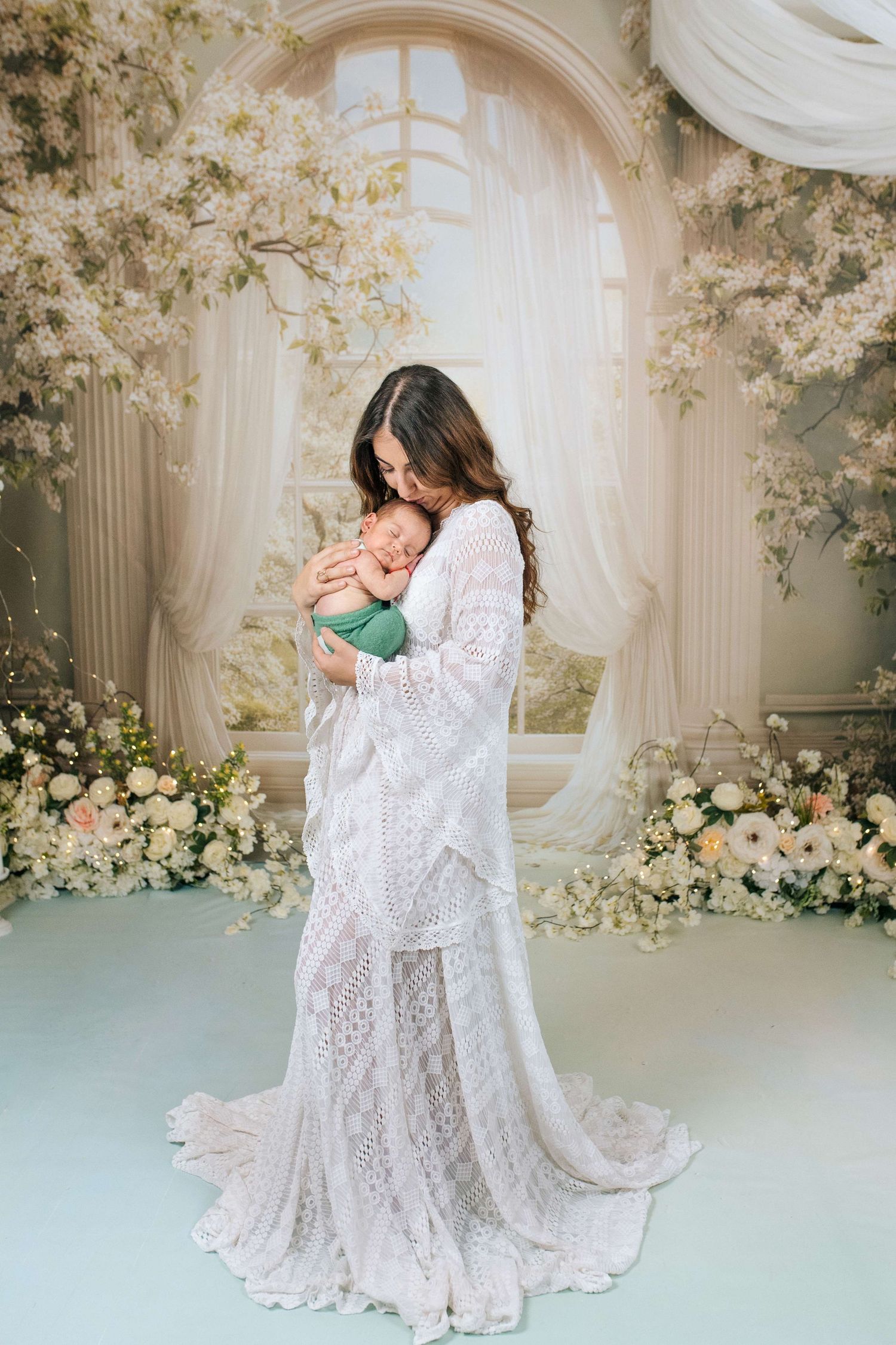 Mother in white lace gown tenderly holds sleeping newborn baby amid elegant floral backdrop with soft draping curtains.