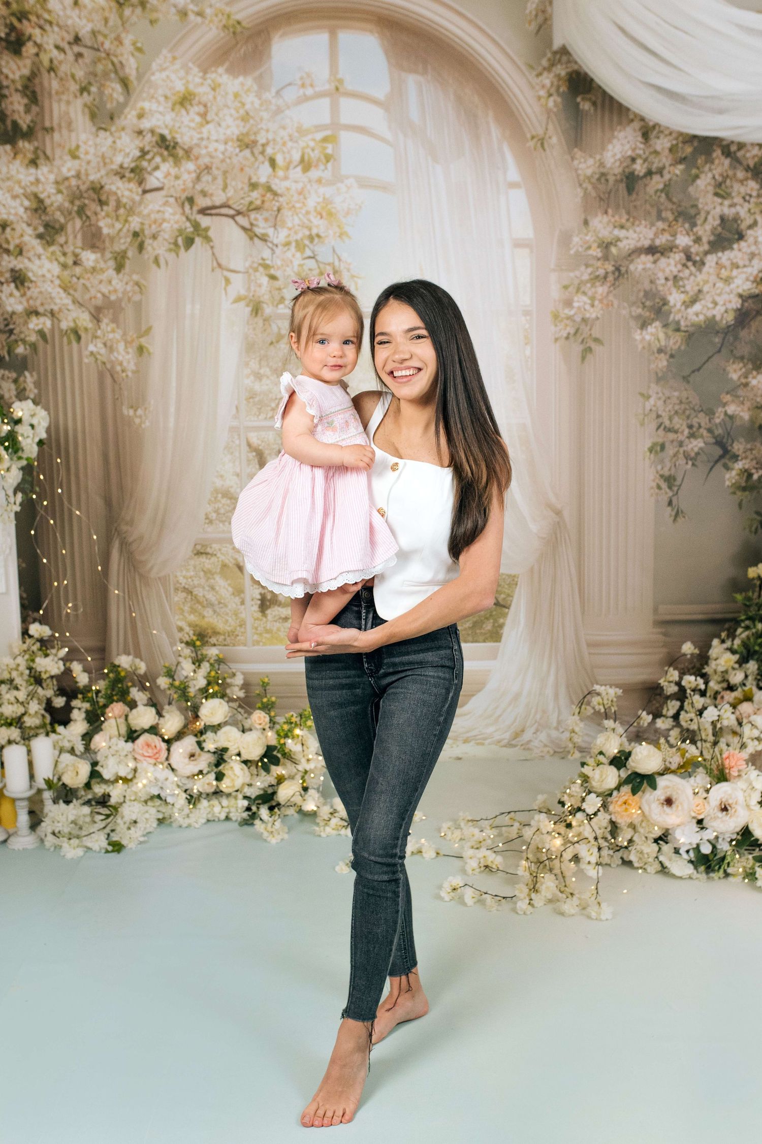 Young woman holding a toddler girl in a pink dress, posing in a floral studio backdrop with white flowers.