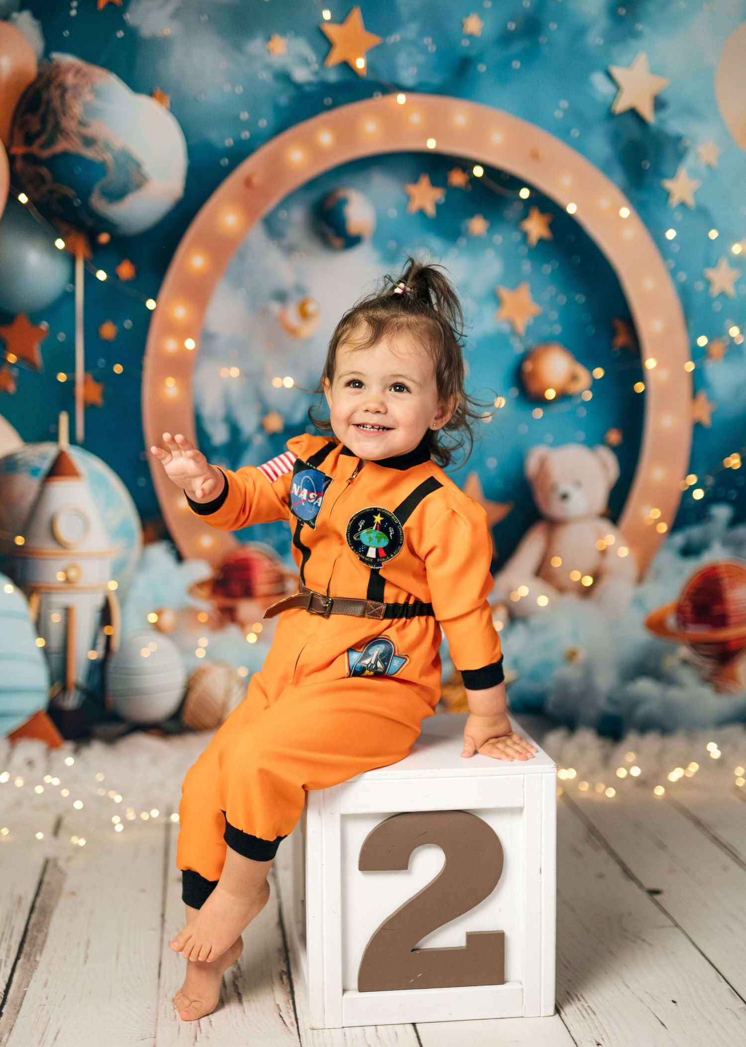 Toddler in orange astronaut suit sits on a number 2 block against a space-themed backdrop with stars and planets.