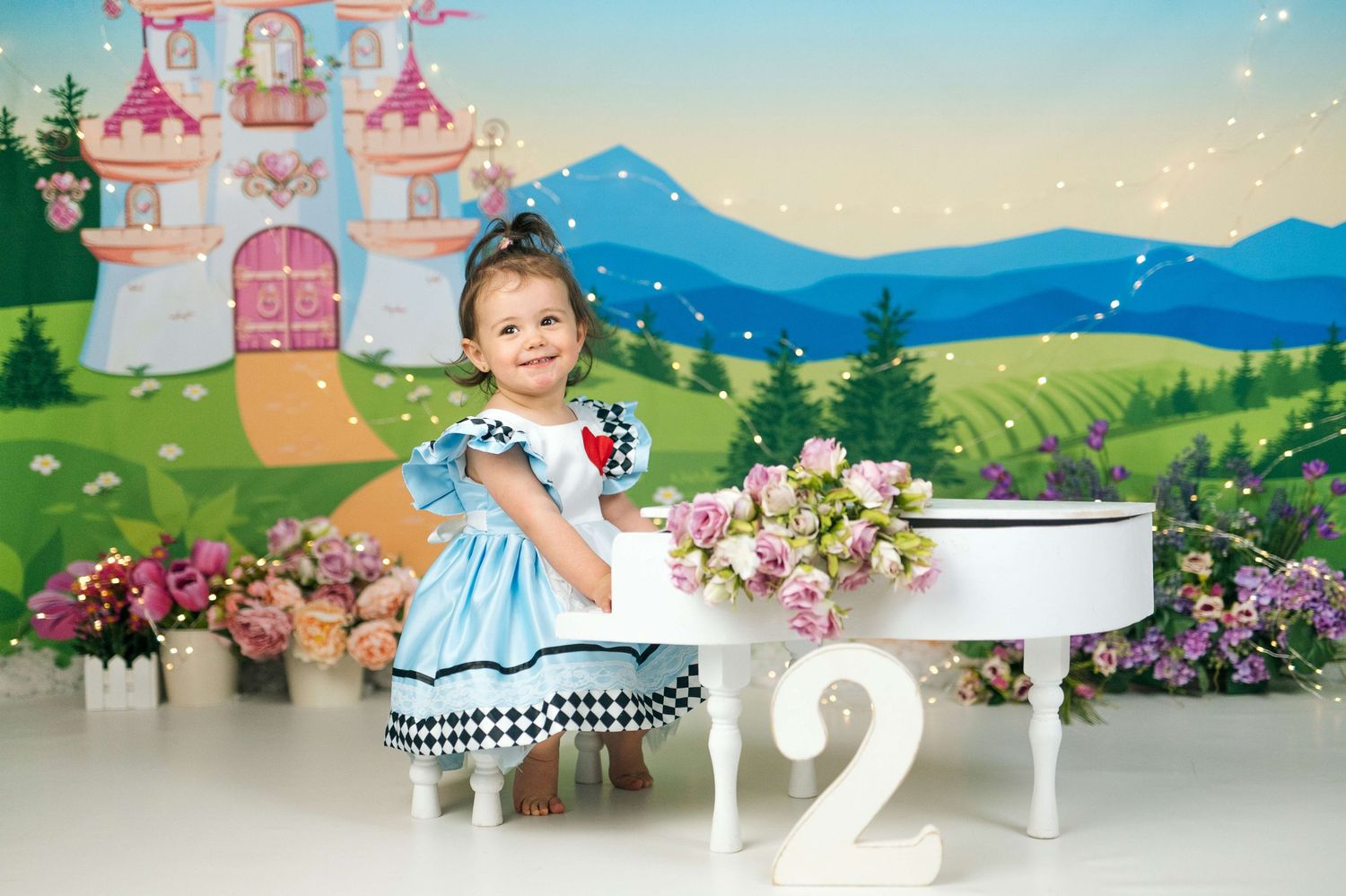 Toddler girl in Alice in Wonderland dress poses by white table with number 2 and flowers against fairy tale castle backdrop.