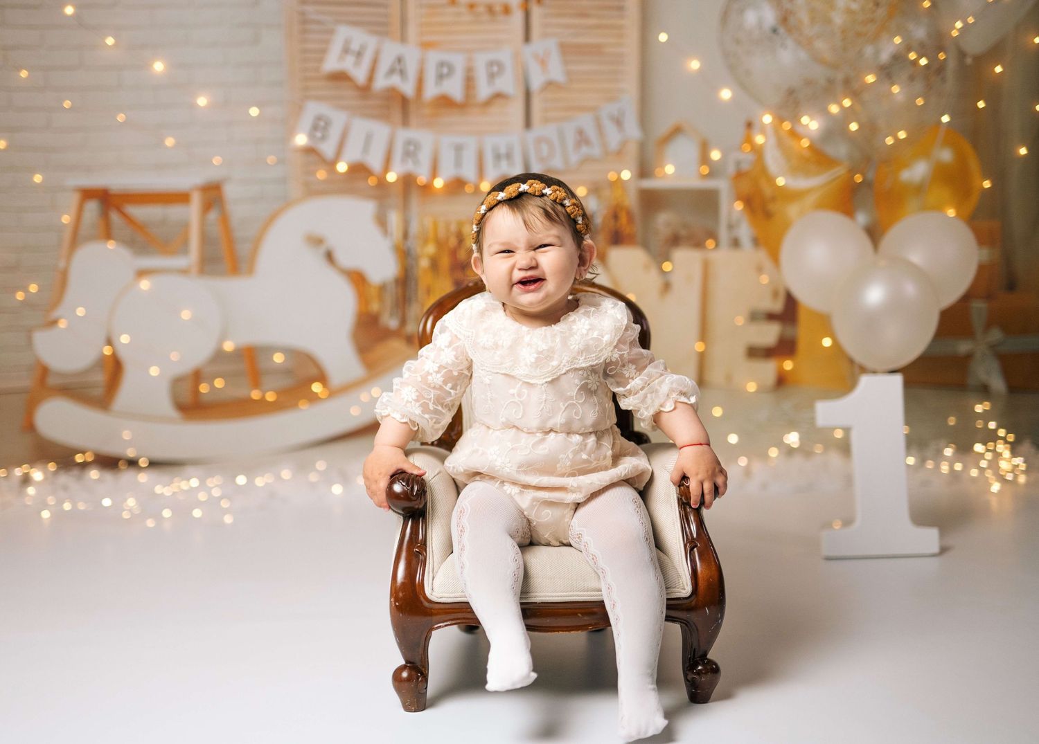 Smiling baby girl in white outfit sitting in chair at first birthday photo shoot with balloons.