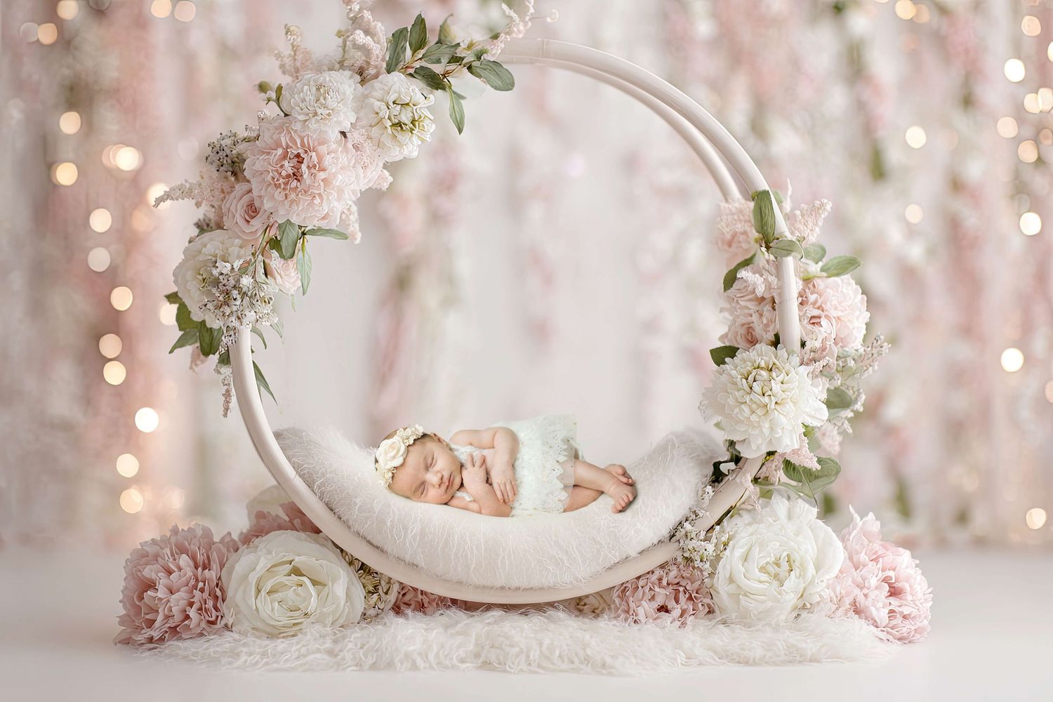 Newborn baby sleeping in a floral wreath with pink and white flowers, soft bokeh lights in the background.