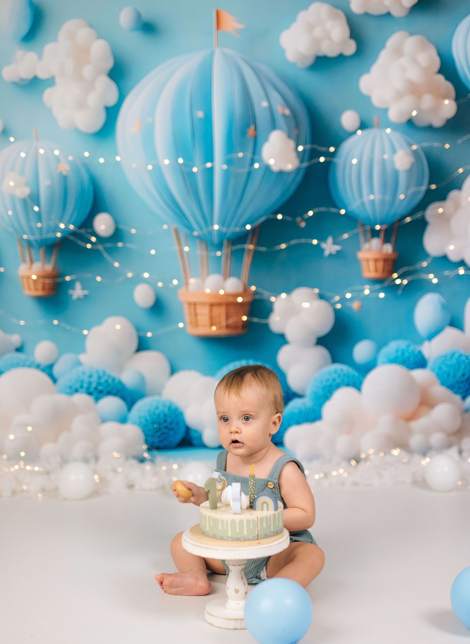 Baby's first birthday cake smash with blue hot air balloon backdrop, white clouds, and fairy lights.