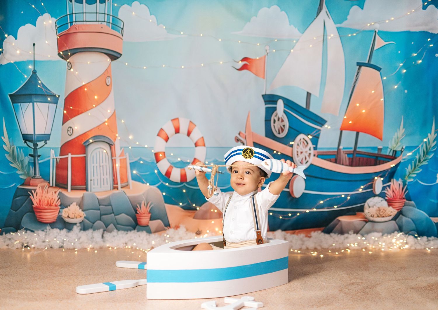 Baby boy in sailor outfit sits in toy boat with nautical backdrop featuring lighthouse and sailboat.