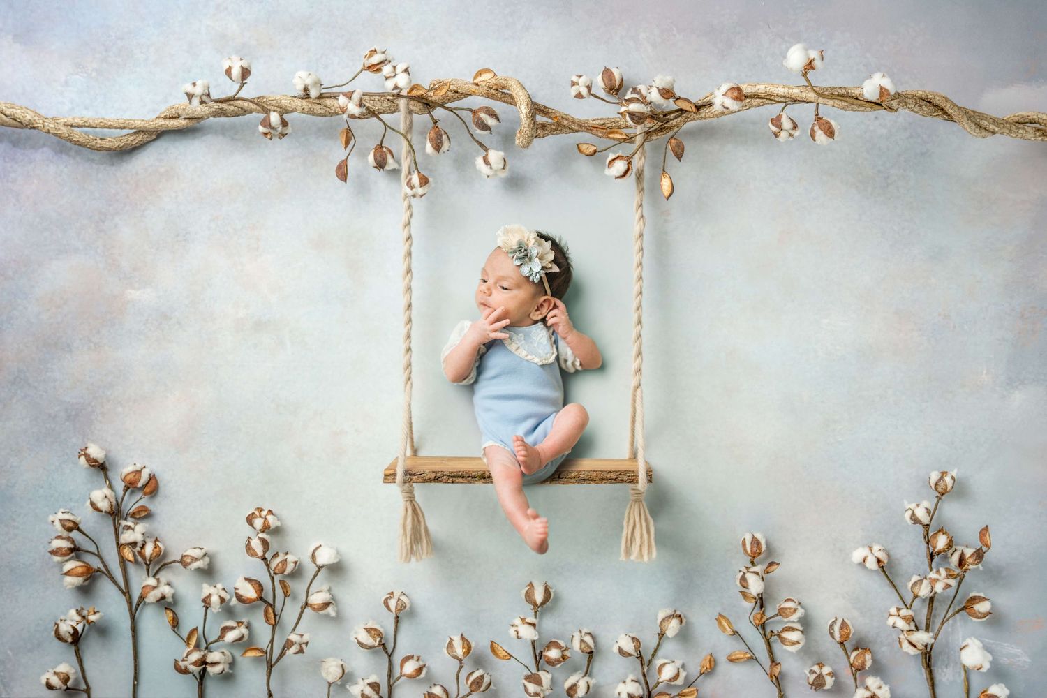 Newborn baby sleeping peacefully on a tiny wooden swing surrounded by cotton flowers on a soft blue background.