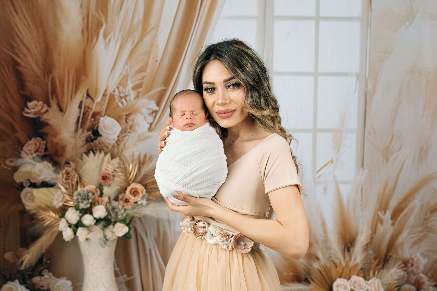 A woman in a beige dress holds a swaddled newborn baby, surrounded by boho floral decor and pampas grass.