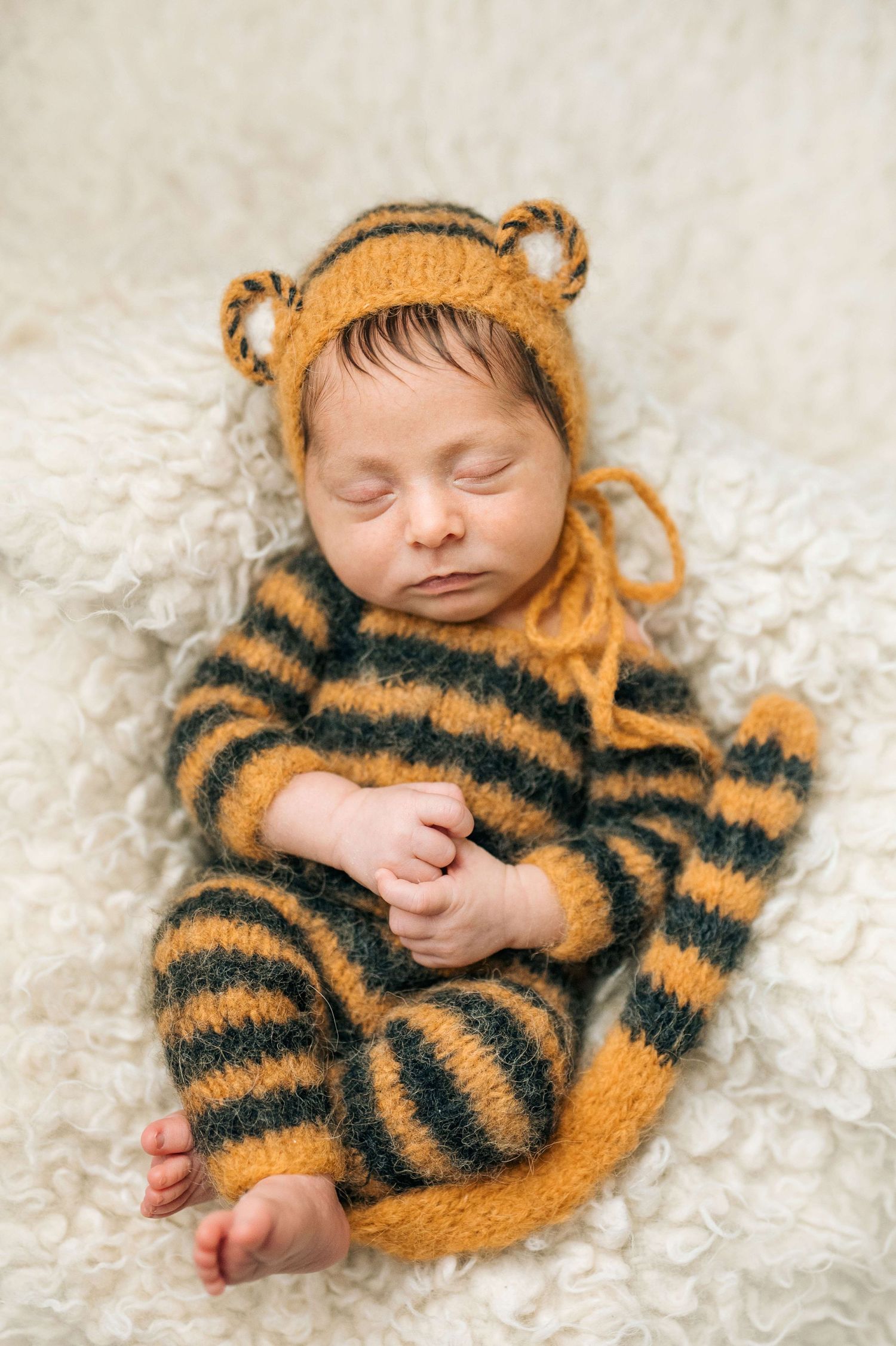 Sleeping newborn baby wearing a knitted yellow and black striped tiger outfit with matching bear ear hat on white fluffy blanket.