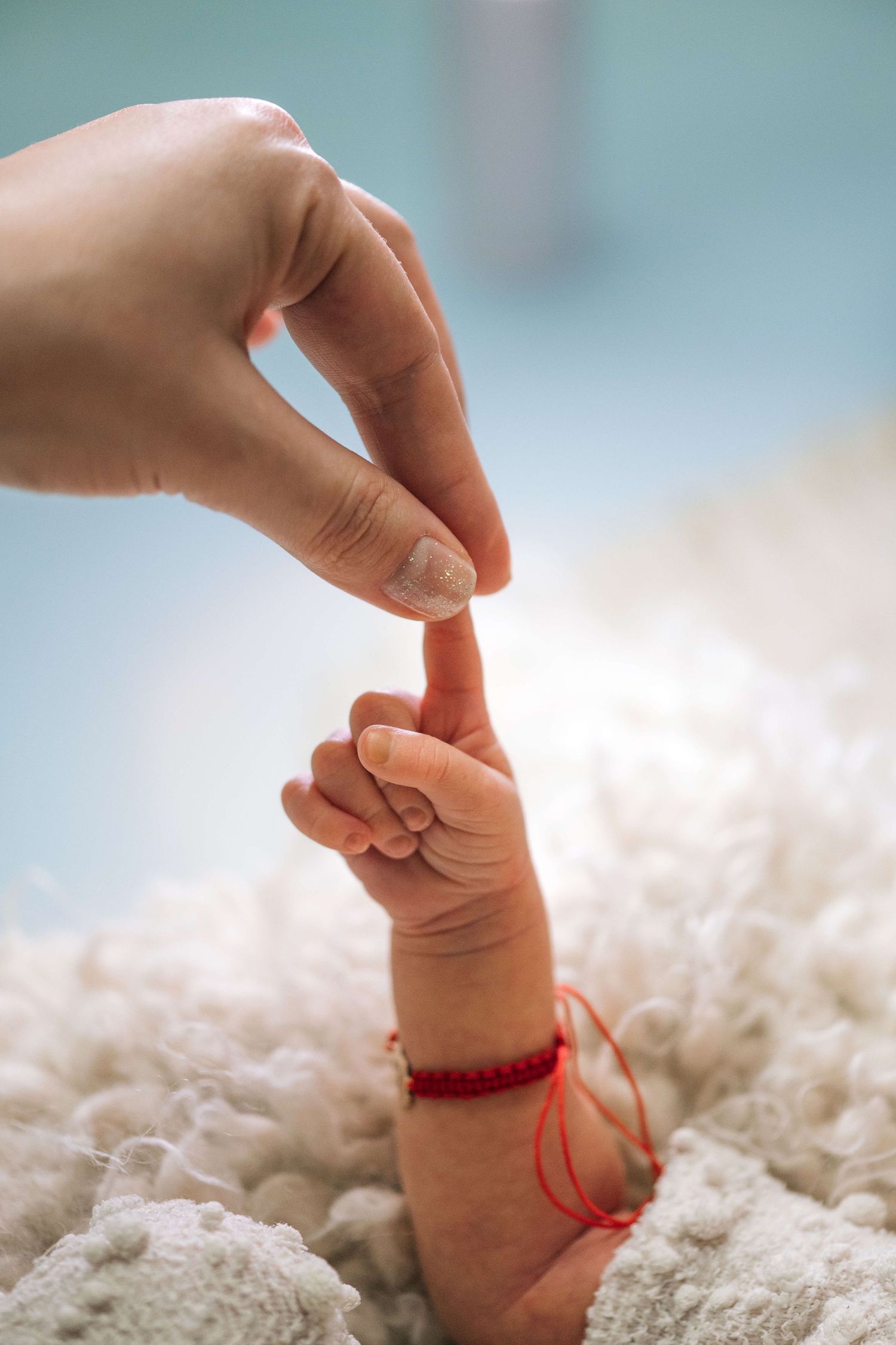 A newborn baby with a red bracelet grasping an adult's finger on a soft white blanket.