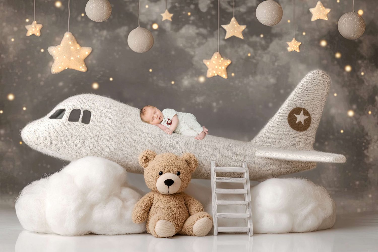 Newborn baby sleeping on a plush toy airplane with a teddy bear, stars, and clouds in a dreamy nursery photo.