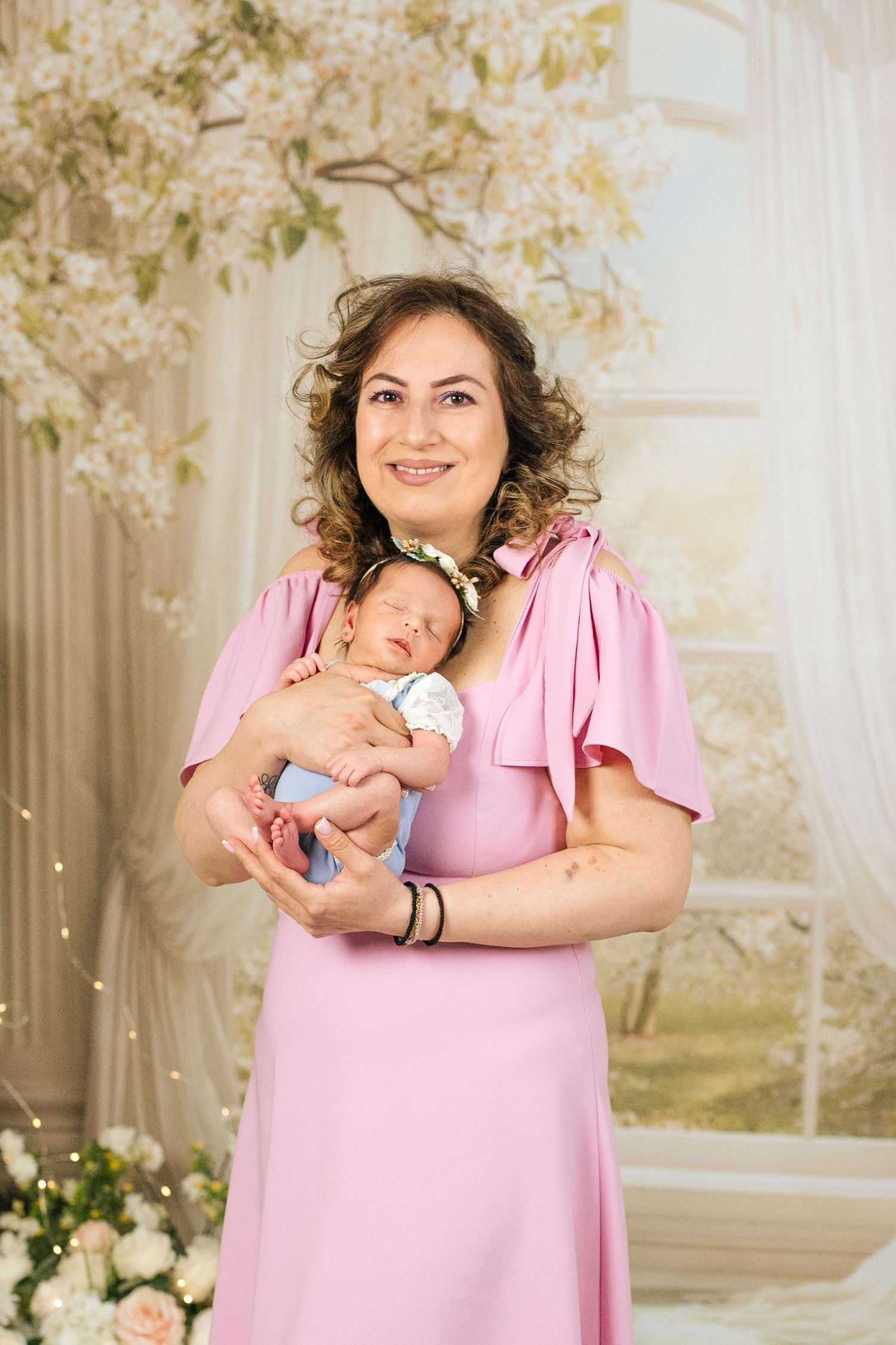 A smiling woman in a pink dress holds a sleeping newborn baby in a floral studio setting.