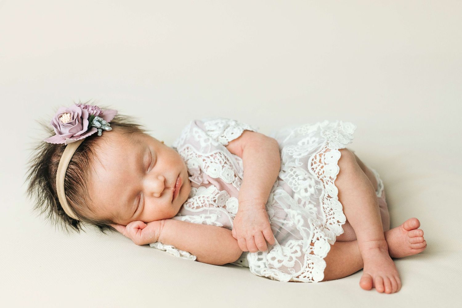 Sleeping newborn baby girl in white lace romper with floral headband on neutral background.