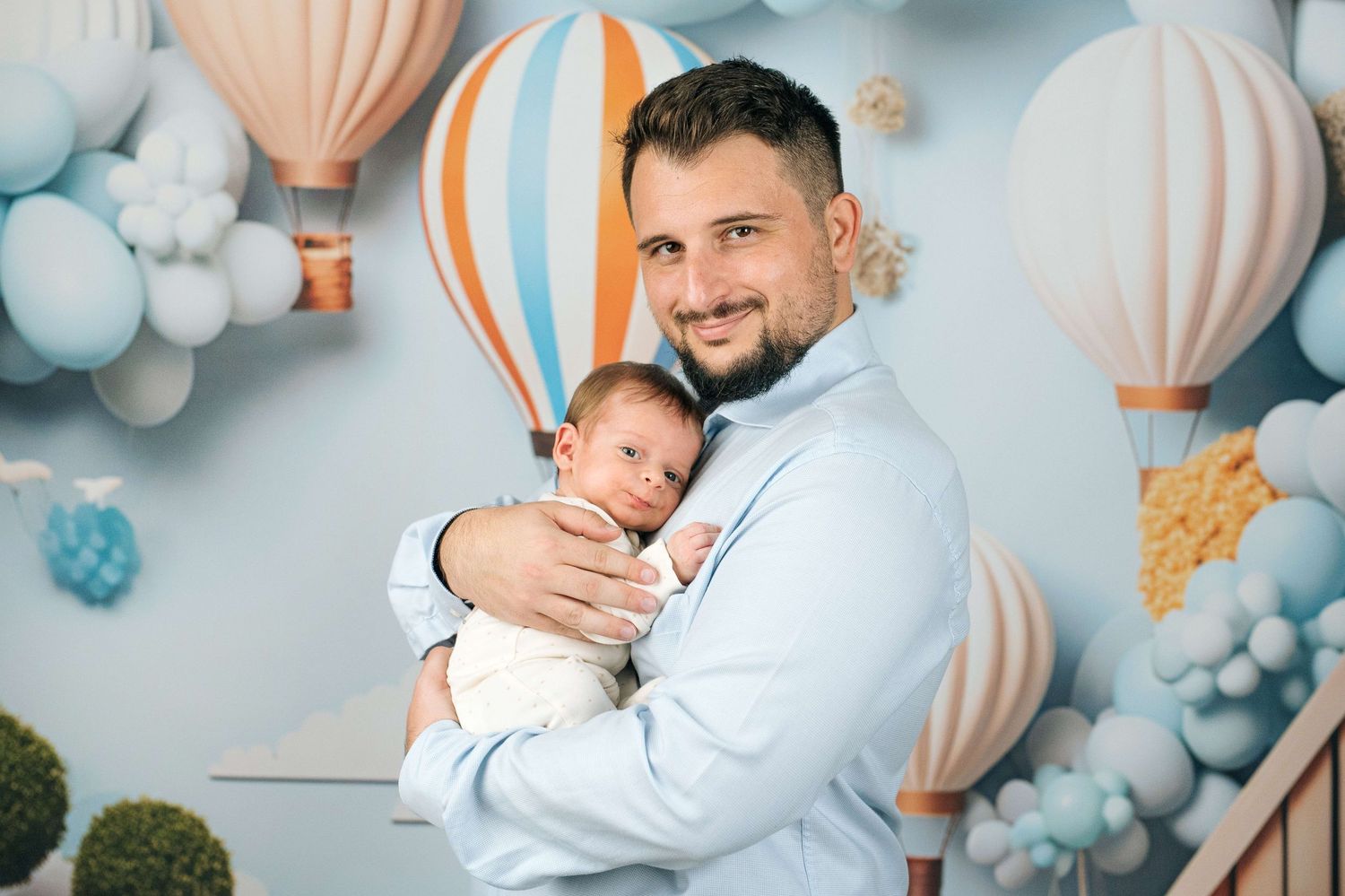 A proud father in a light blue shirt holds his newborn baby against a whimsical hot air balloon backdrop.