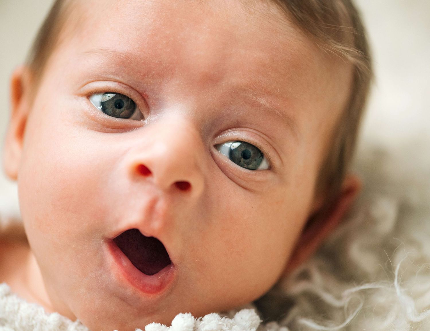 Close-up portrait of a wide-eyed newborn baby with blue eyes and open mouth on a soft blanket.