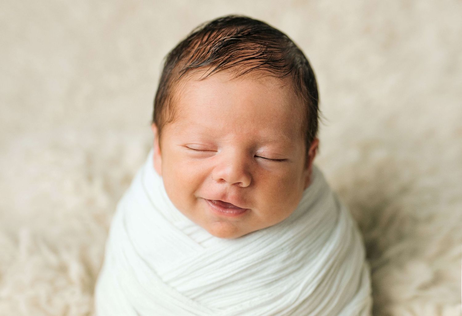 Smiling newborn baby swaddled in white wrap sleeping peacefully on a soft cream blanket.