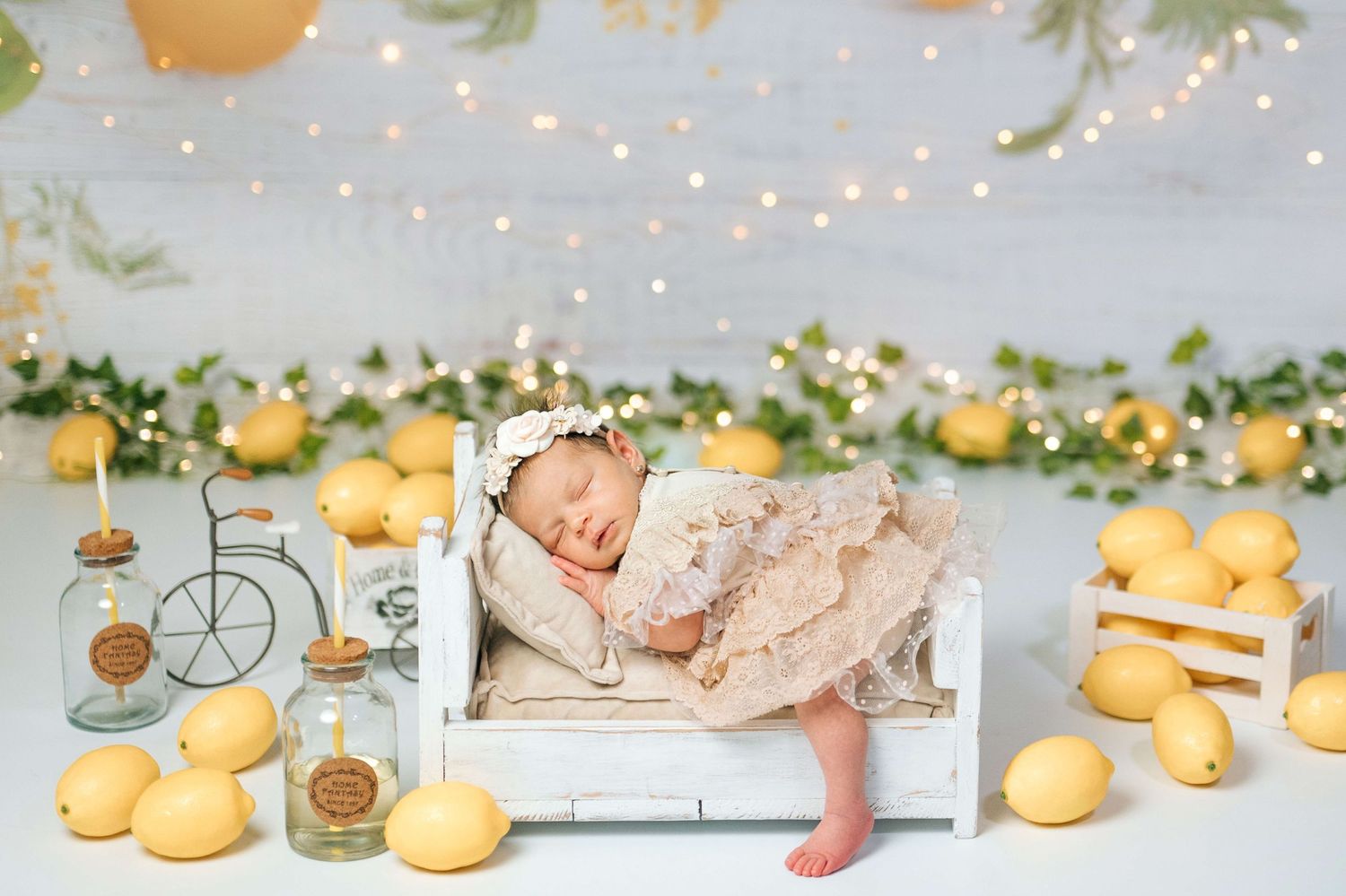 Newborn baby sleeping on tiny white bed surrounded by lemons, fairy lights, and greenery in dreamy studio photo.