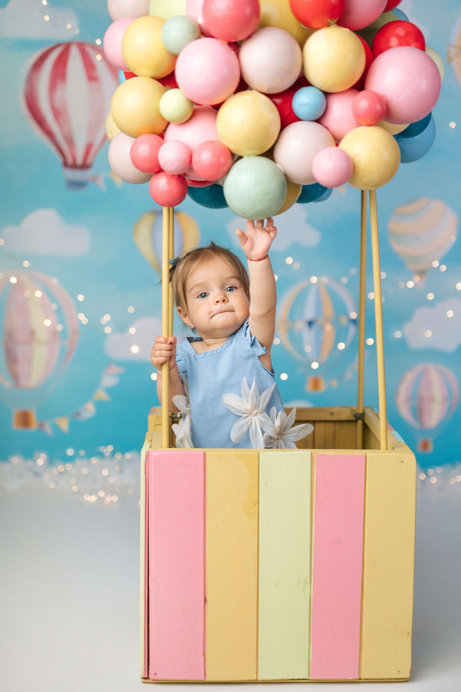 Baby in blue outfit sitting in colorful striped hot air balloon basket with pastel balloons against sky backdrop.