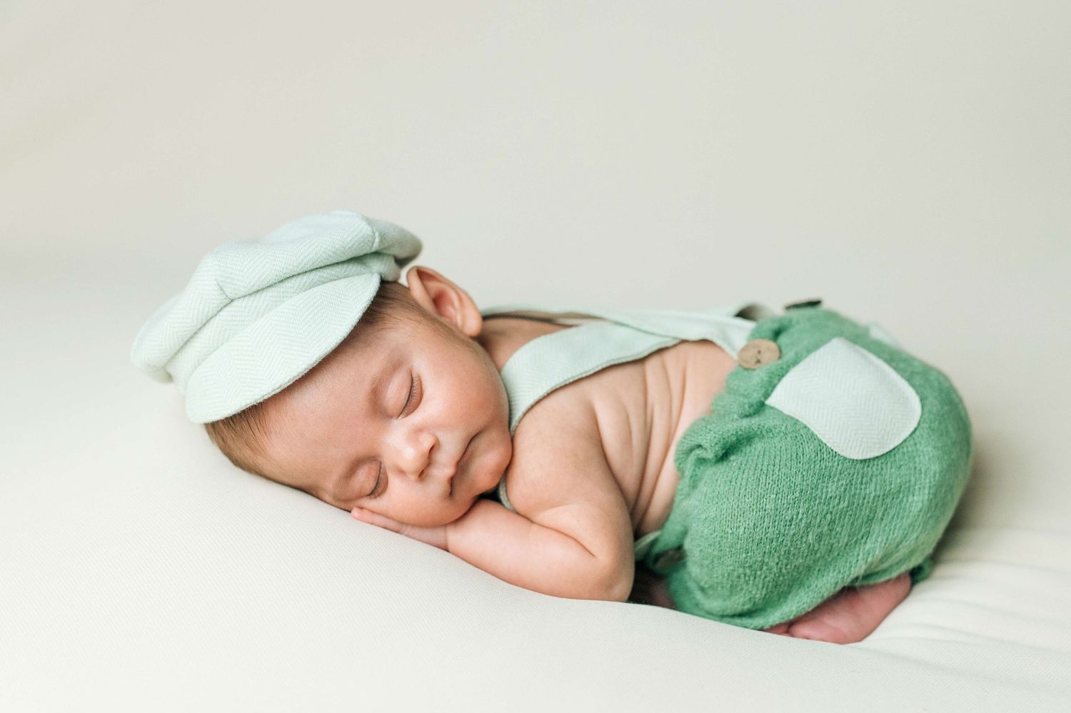 Sleeping newborn baby wearing a green frog costume with hat, posed on a white blanket.