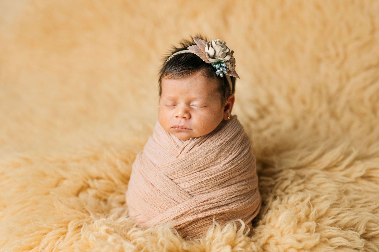 Newborn baby girl swaddled in pink wrap with floral headband sleeping on fluffy beige fur backdrop.