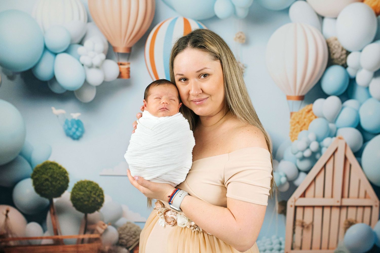 Mother holding swaddled newborn baby in front of blue balloon and hot air balloon themed backdrop.