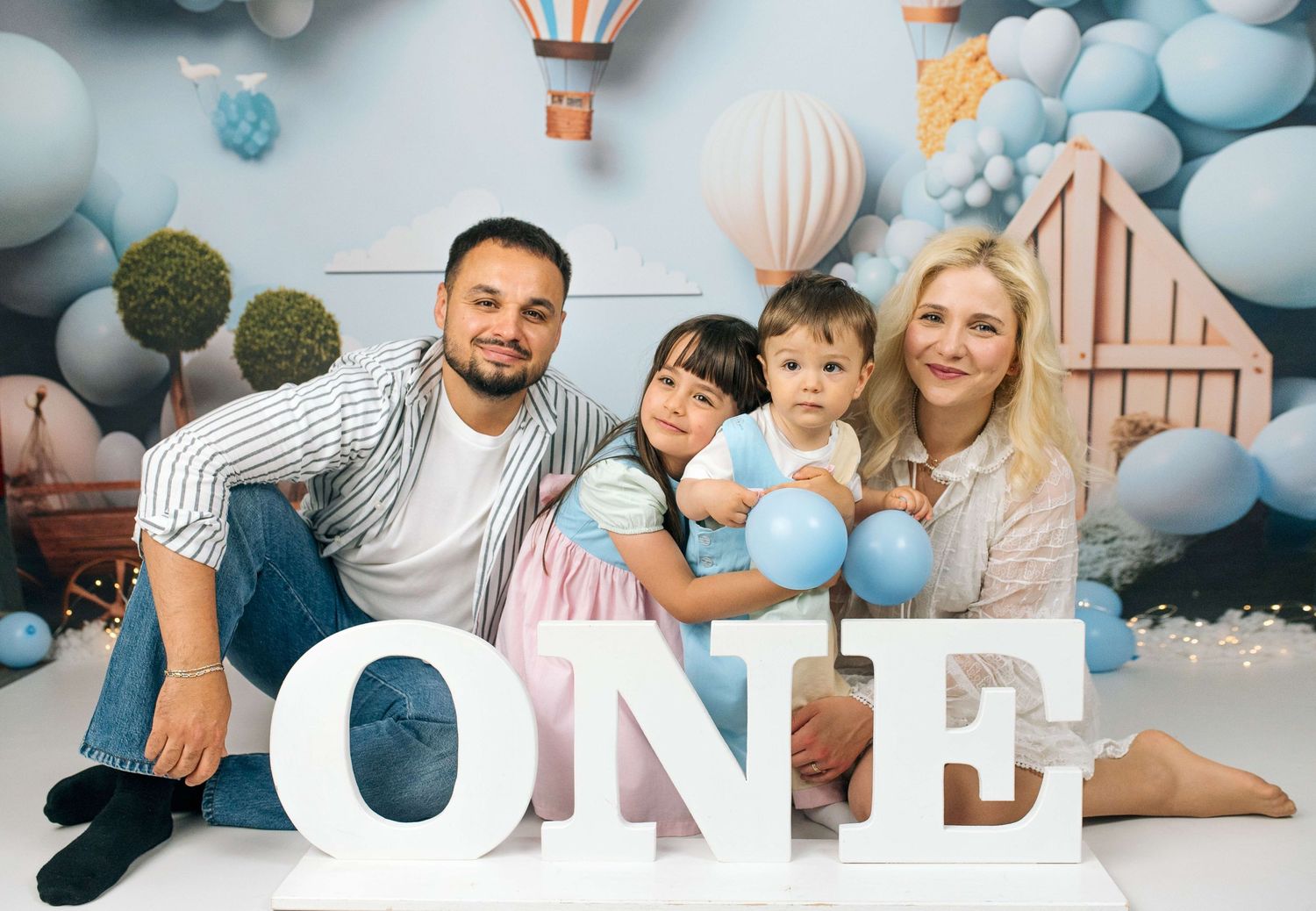 Family of four posing with ONE letters at a baby's first birthday party with blue balloon decorations.