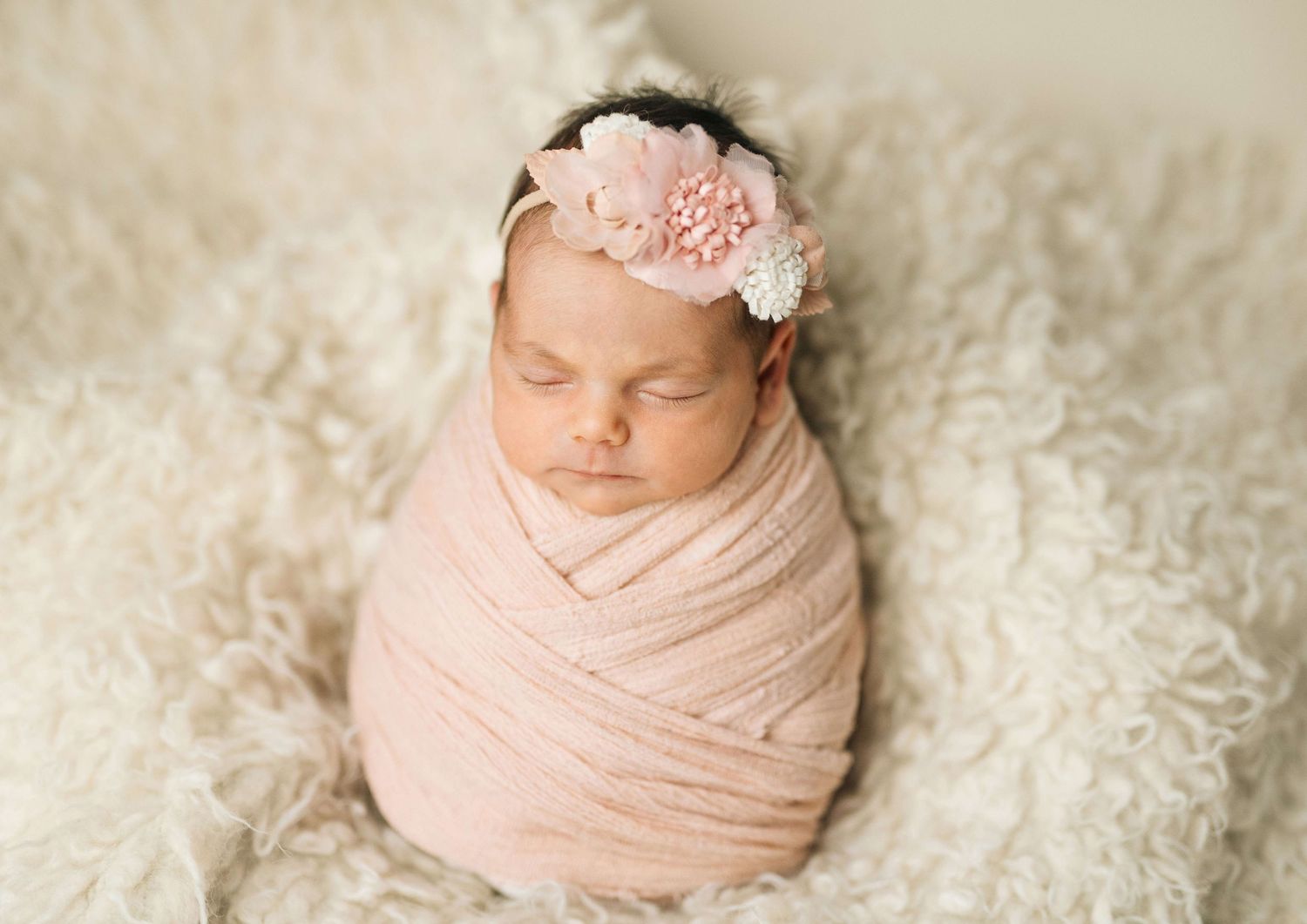 Newborn baby girl swaddled in pink wrap with floral headband, sleeping peacefully on white fluffy blanket.