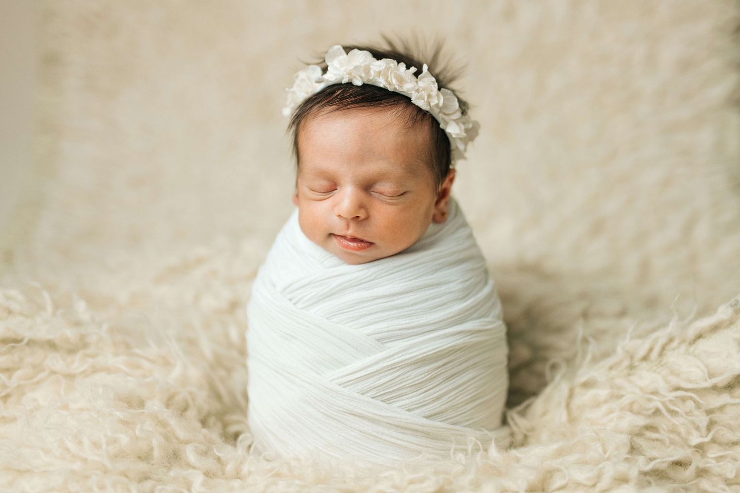 Sleeping newborn baby swaddled in white wrap wearing floral headband on fluffy cream backdrop.
