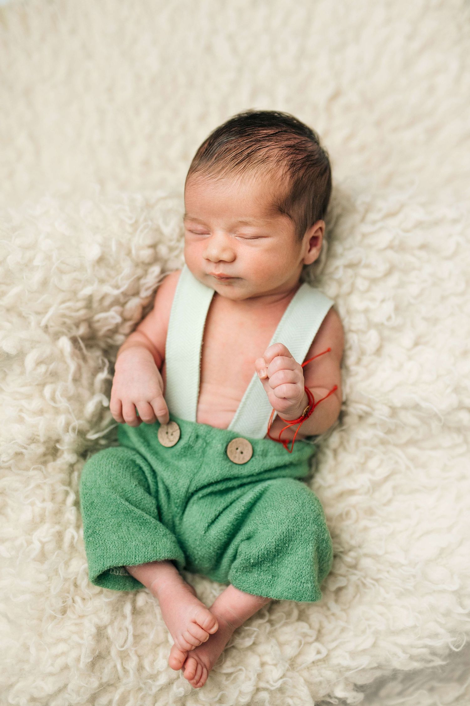 Sleeping newborn baby wearing green suspender pants lying on a fluffy cream blanket.