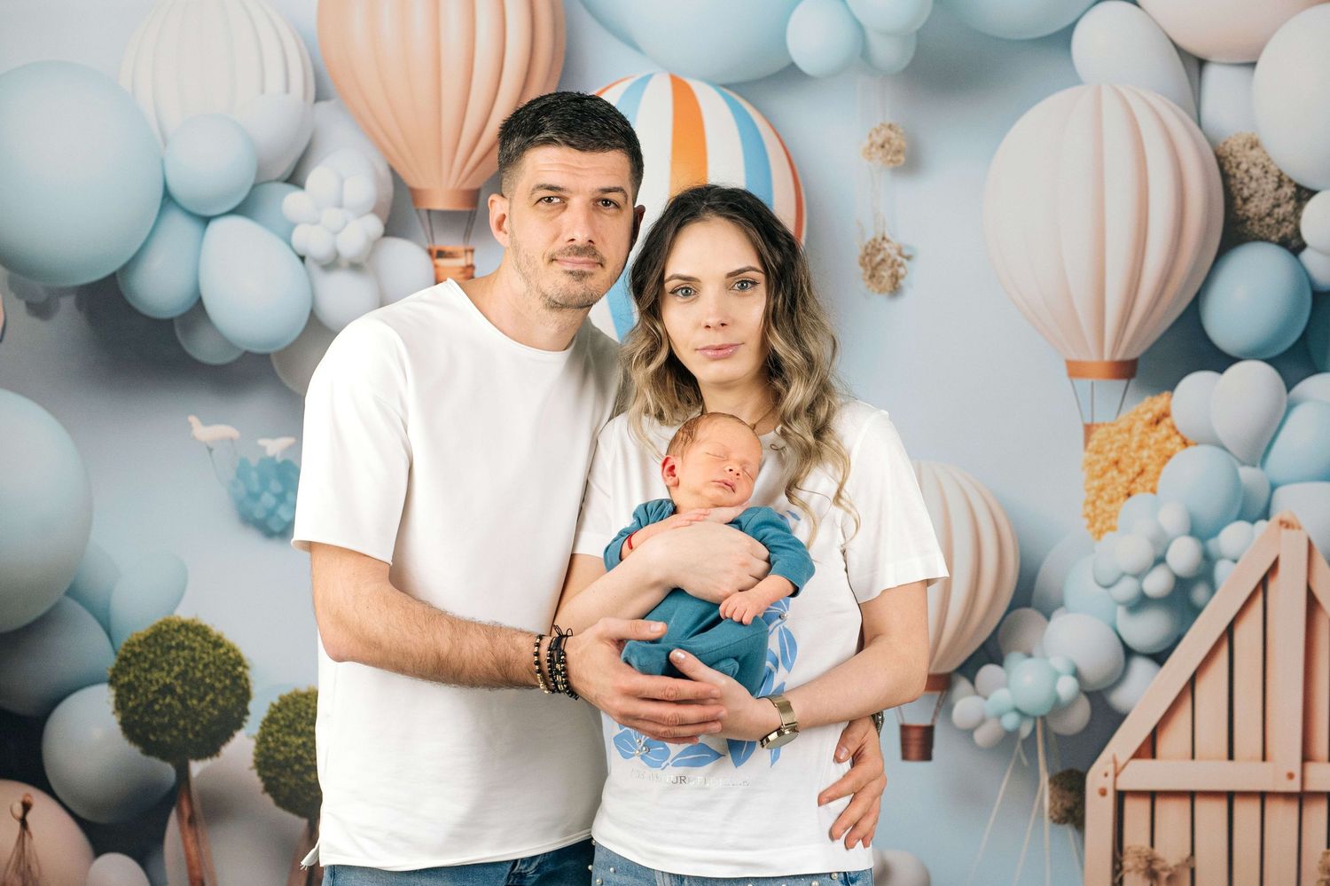 A young couple in white t-shirts holds their newborn baby in front of a decorative hot air balloon backdrop.