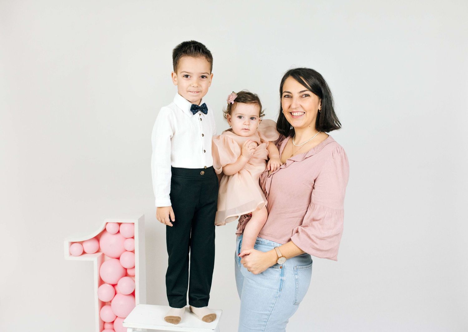 A woman holds a baby girl while a young boy stands beside a pink number one decoration for a first birthday photo.