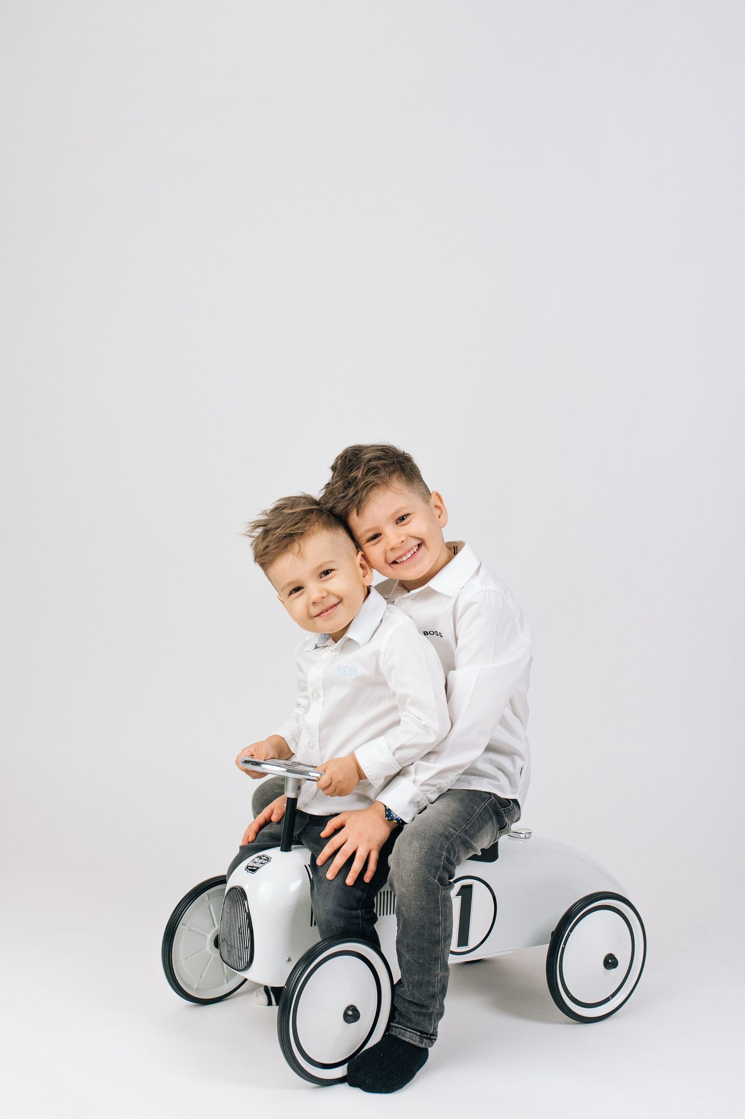 Two smiling boys in white shirts sit together on a white toy ride-on car against a light gray background.