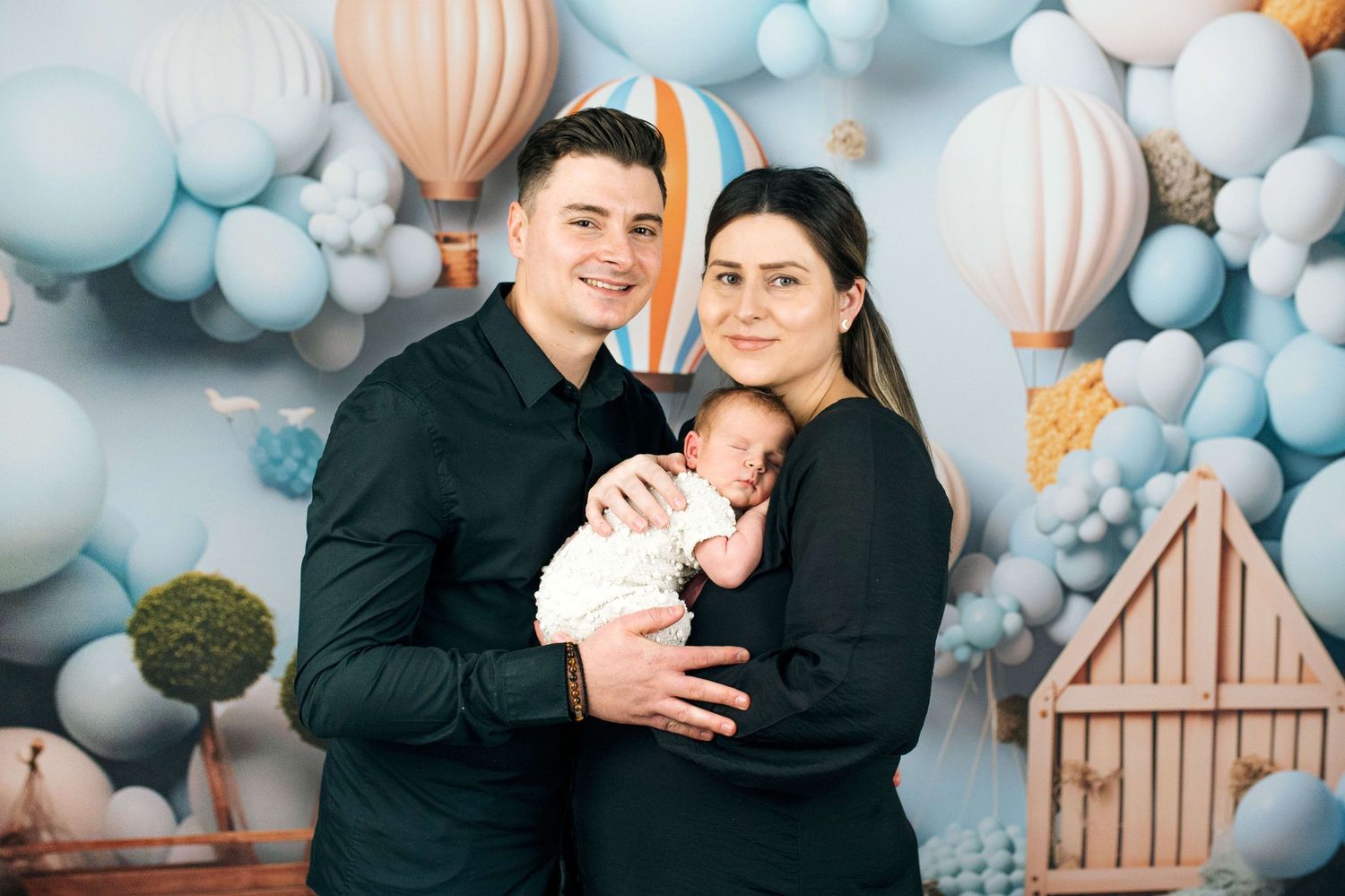 Young couple in black outfits holding newborn baby in front of blue balloon and hot air balloon backdrop.