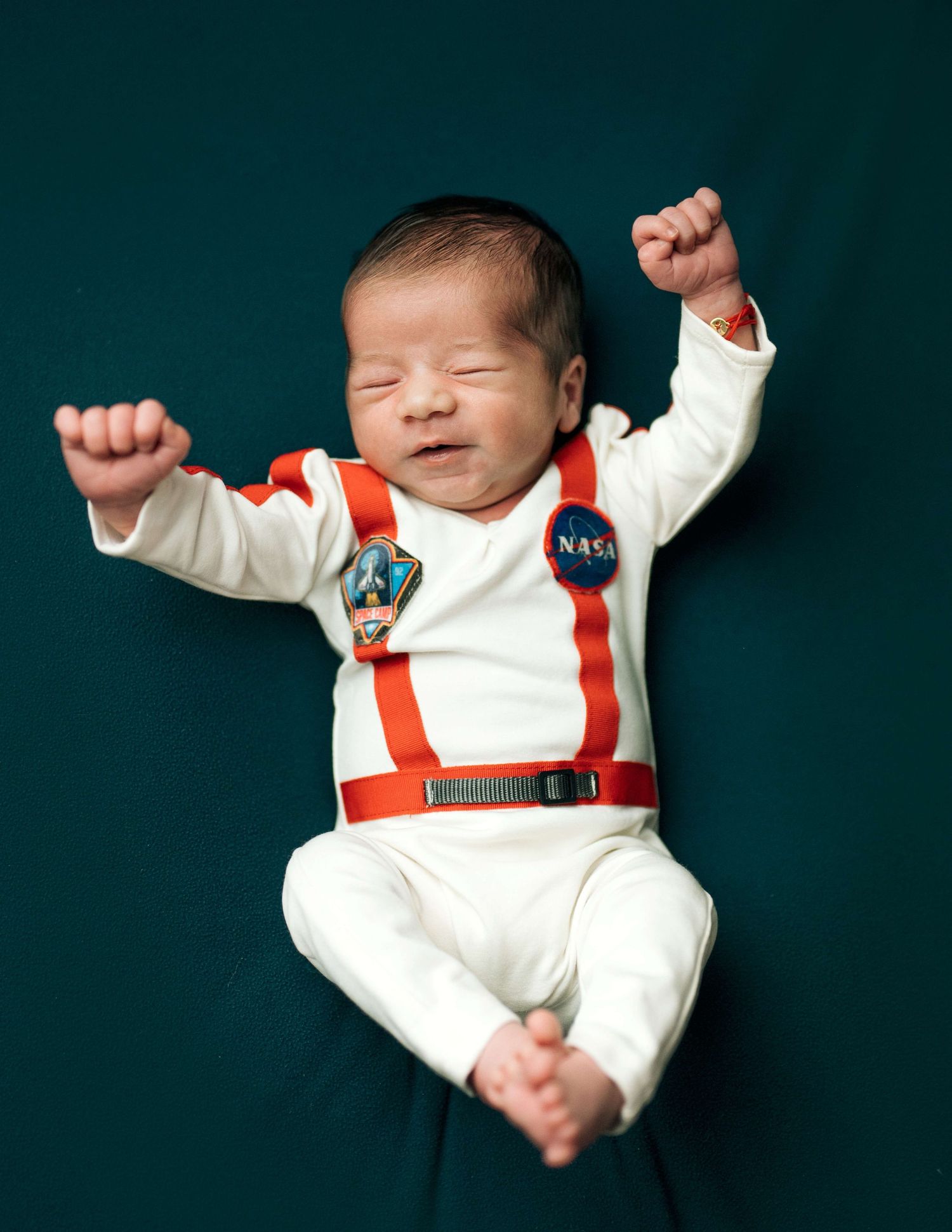 Newborn baby in a white NASA astronaut costume with patches and suspenders, arms raised, lying on teal background.