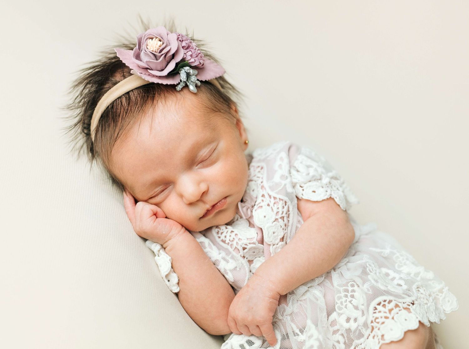 Sleeping newborn baby girl in white lace outfit and purple floral headband, posed peacefully on cream background.