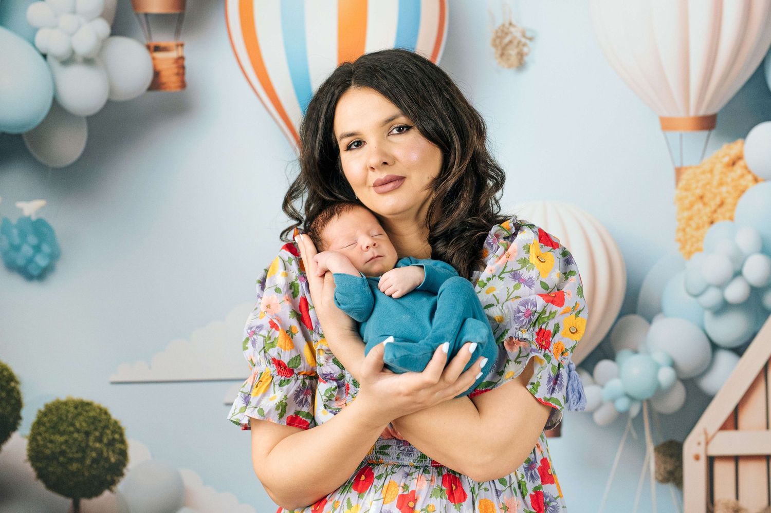 Woman in floral dress holds sleeping newborn in blue outfit against decorative balloon backdrop.