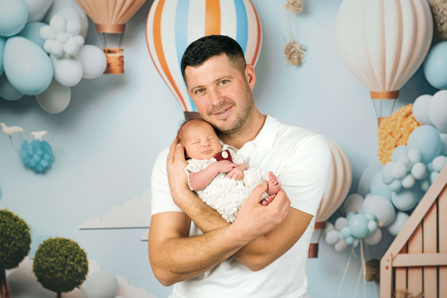 Father holding newborn baby in front of blue balloon backdrop with hot air balloons during professional photo shoot.