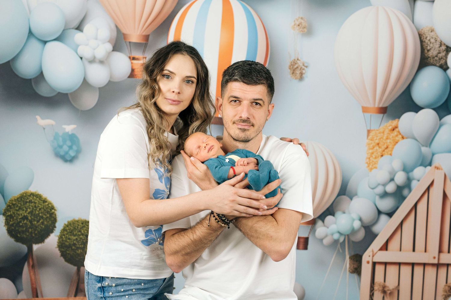 Young couple in white t-shirts holding newborn baby in blue wrap, posing before hot air balloon backdrop.