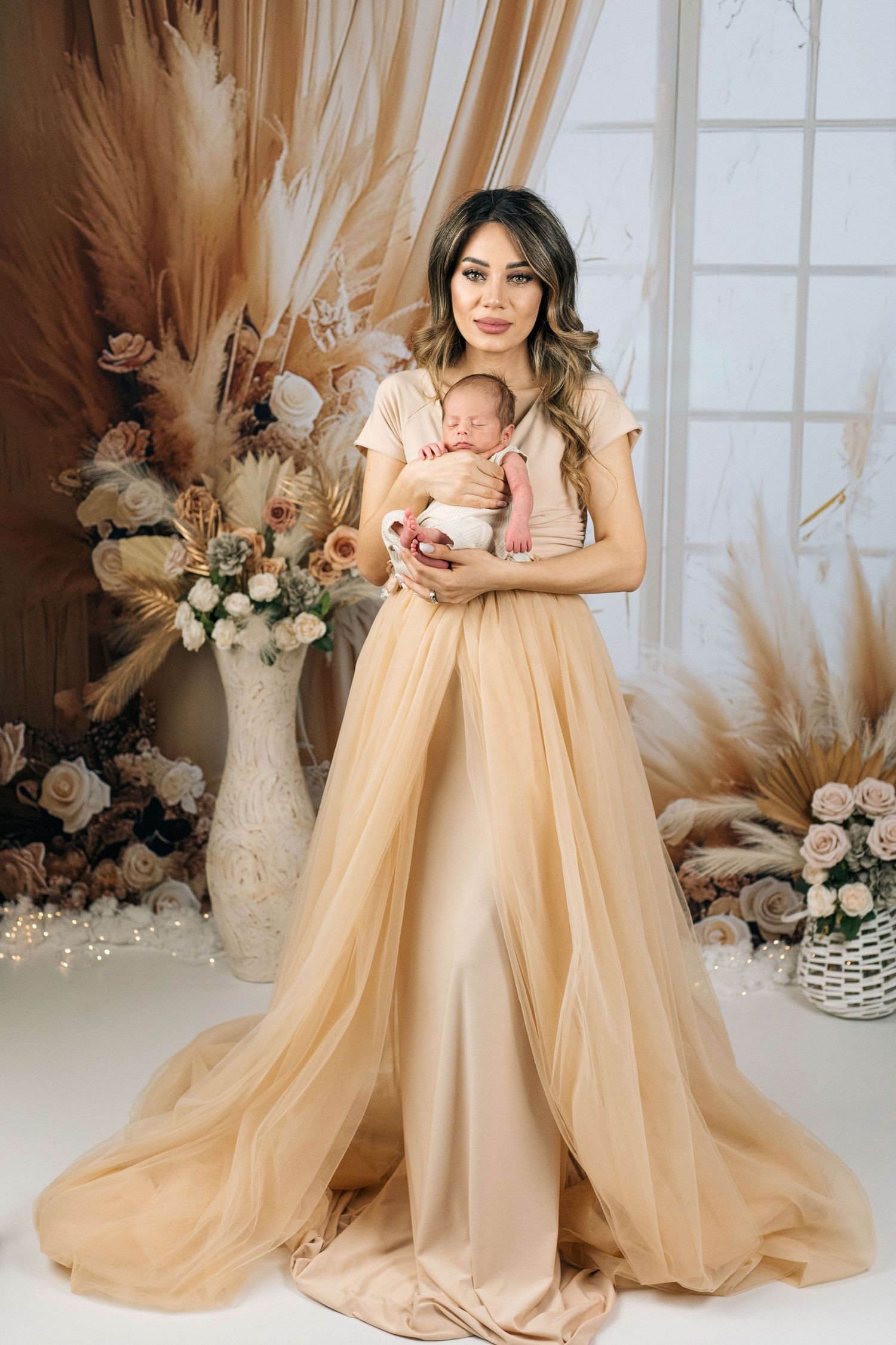 Elegant woman in gold tulle gown holding newborn baby, surrounded by pampas grass and floral arrangements.