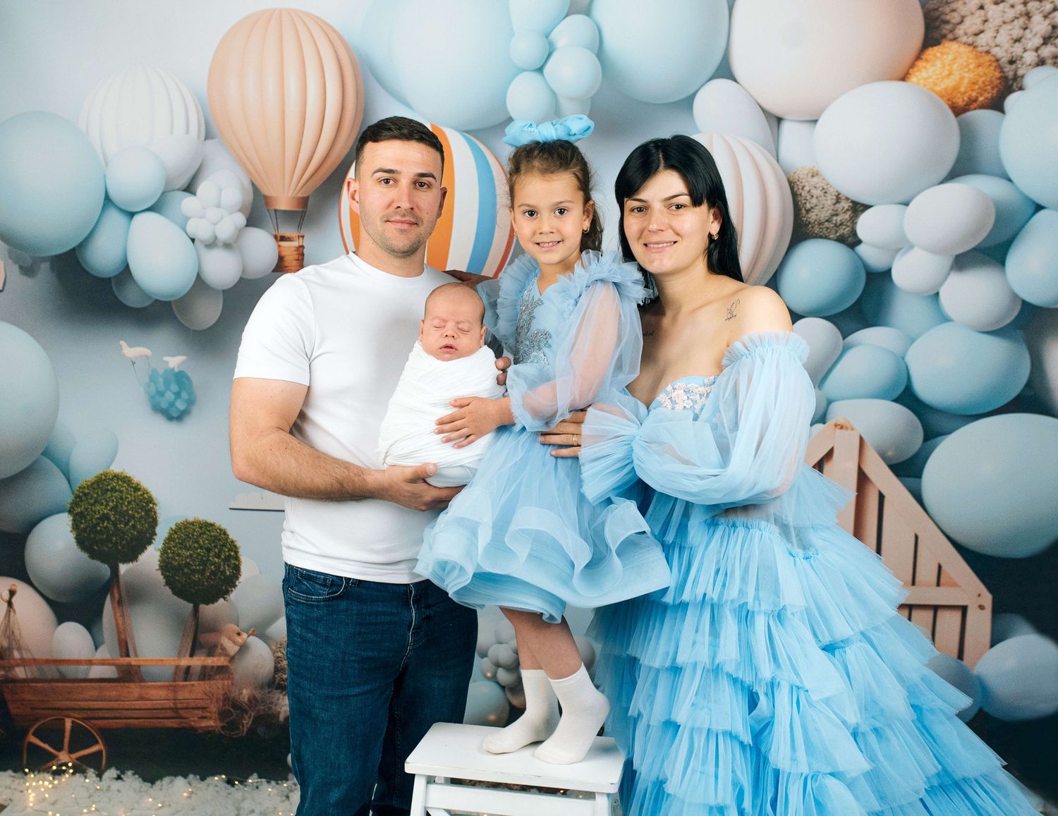 Happy family of four posing with blue balloons backdrop, parents holding newborn and toddler girl in matching blue outfits.