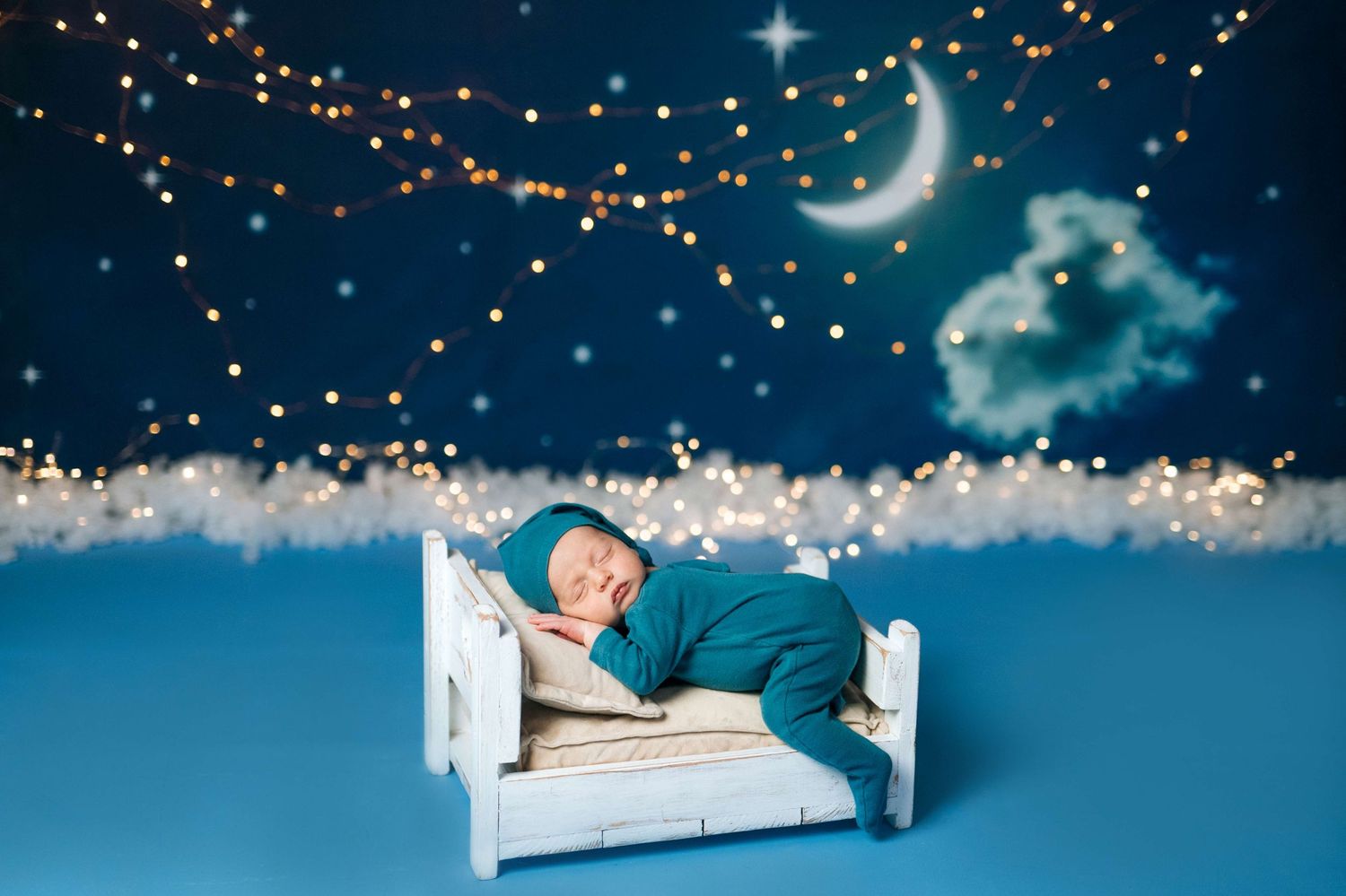 Newborn baby sleeping peacefully in tiny white bed with teal outfit, crescent moon and fairy lights backdrop.