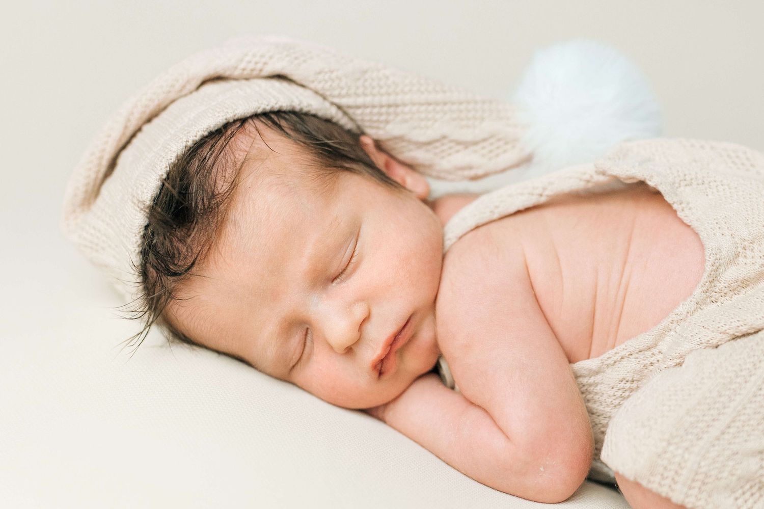Sleeping newborn baby wearing a knit bunny hat with blue pompom tail in a cozy beige outfit.