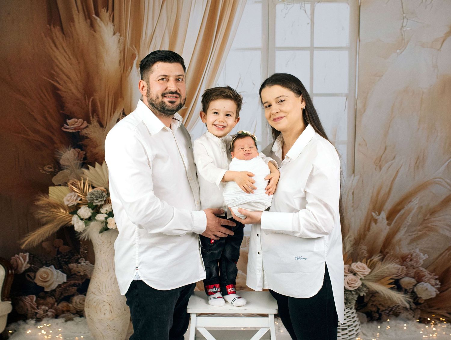 Family portrait with parents and two children, including a newborn baby, posing in a elegant floral studio setting.