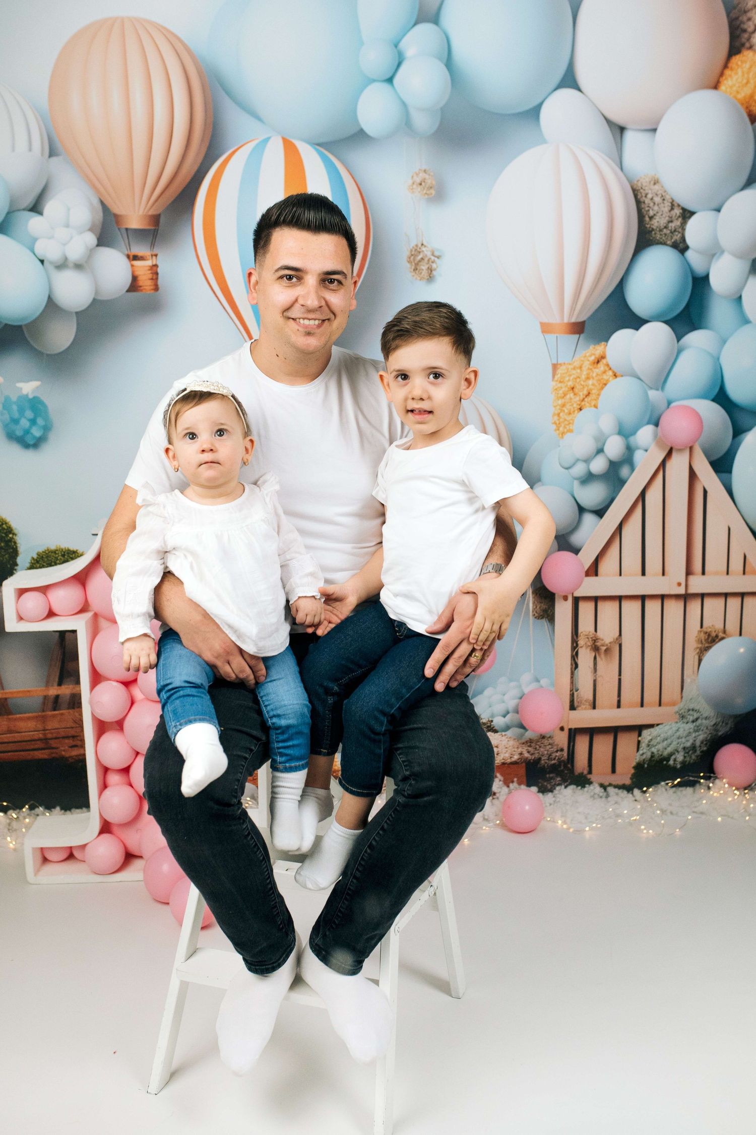 Father sits with two young children in front of colorful hot air balloon backdrop for a first birthday photo shoot.