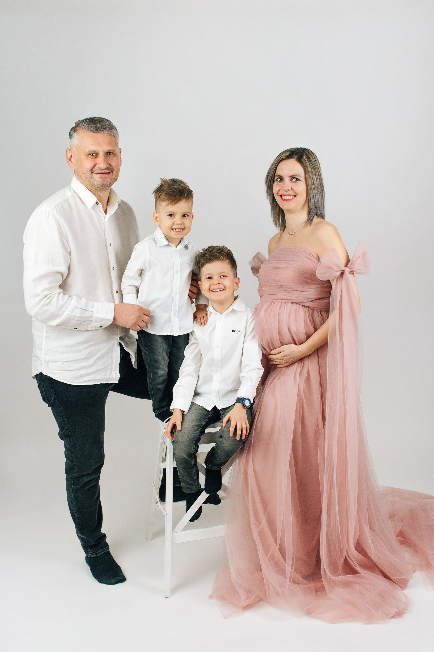 Pregnant woman in pink gown poses with husband and two young sons in white shirts for a family studio portrait.