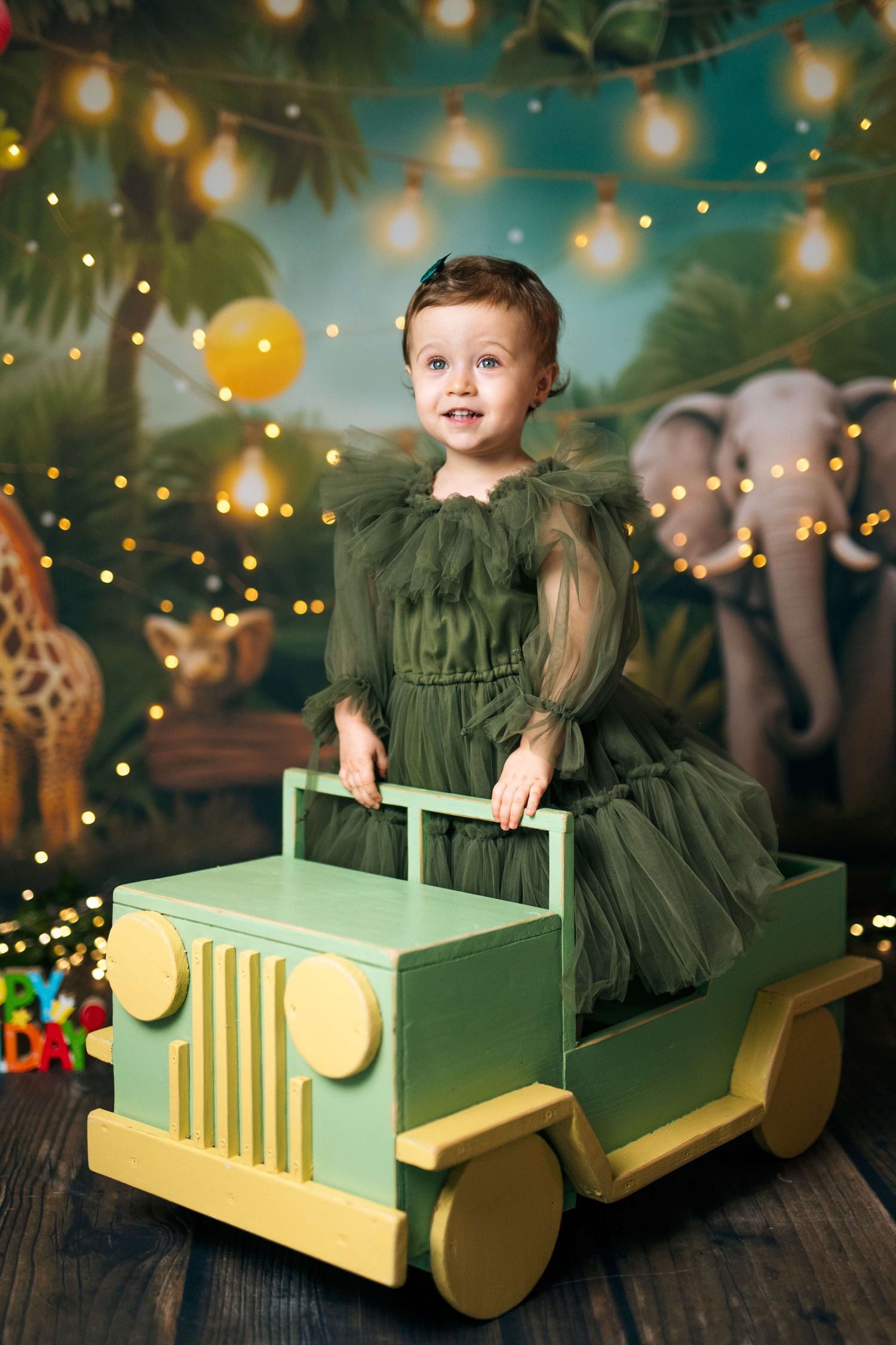 Toddler girl in green tulle dress sitting in wooden toy jeep against jungle safari backdrop with string lights.