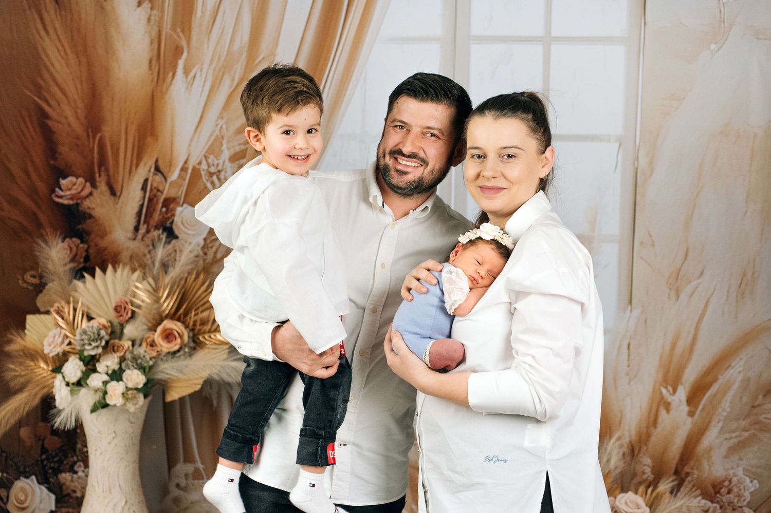 Happy family of four posing indoors, parents holding toddler and newborn baby, wearing white outfits with floral decor.