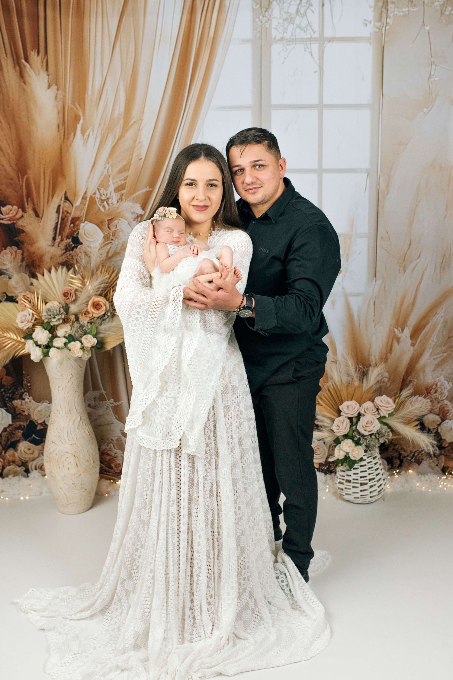 A couple in elegant attire holds their newborn baby surrounded by boho floral arrangements in a studio portrait.