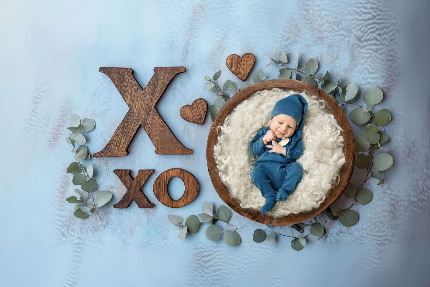 Newborn baby in blue outfit sleeping in wooden bowl surrounded by eucalyptus leaves and XO wooden letters on blue background.