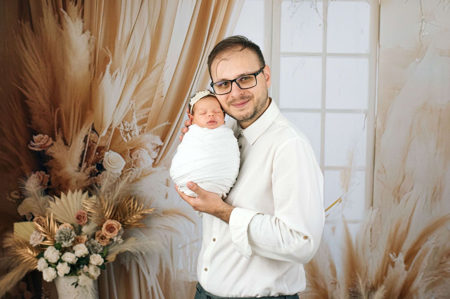 A smiling man wearing glasses holds a swaddled newborn baby in a warmly decorated studio with dried floral arrangements.