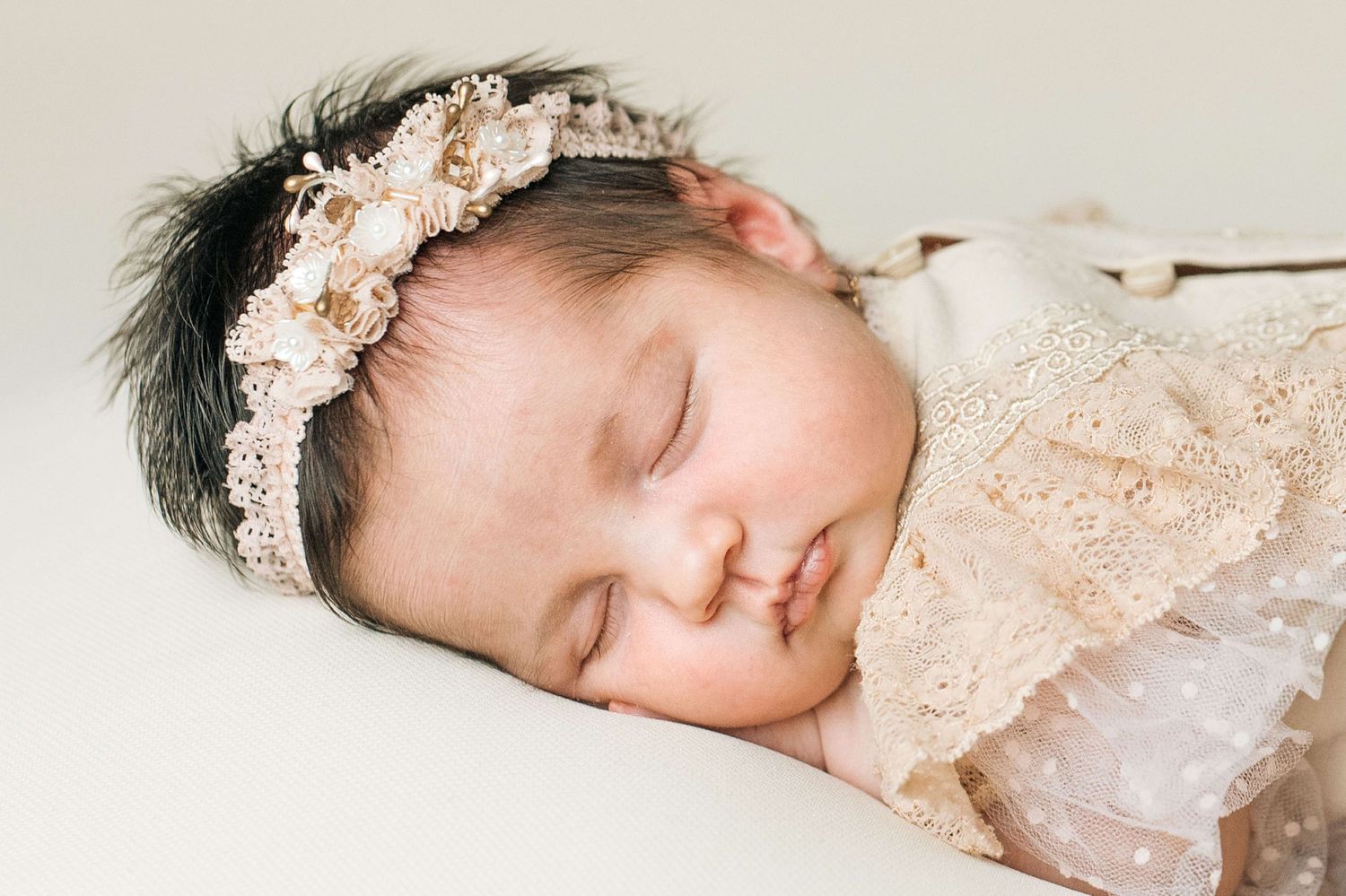Sleeping newborn baby girl wearing a delicate lace headband with pearls and flowers, wrapped in soft pink lace.